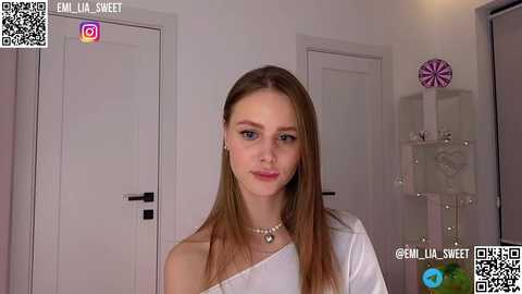 Media: Video of a young Caucasian woman with straight, light brown hair, wearing a white off-shoulder top, standing in a modern, minimalist room with white walls and glass shelving.