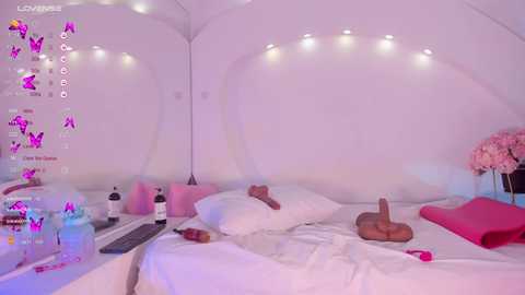 Media: A video of a minimalist, pink-themed bedroom with a white bed, pink pillows, and a sex toy on a pink blanket. The room is illuminated by fairy lights and has a serene, sensual ambiance.