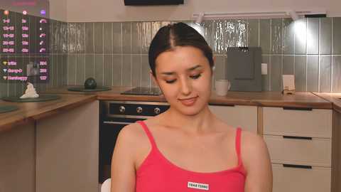 Media: Video of a young Asian woman with straight black hair in a pink tank top, sitting in a modern kitchen with green tiles, wooden countertops, and appliances.