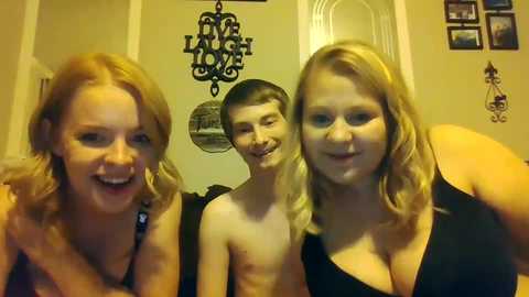 Media: Video of three smiling people: a blonde woman with large breasts in a black top, a shirtless young boy, and a blonde woman with large breasts in a black top, standing in a warmly lit room with decorative wall hangings.