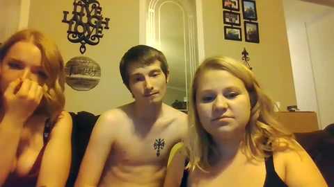 Media: Video of two shirtless teenage boys and a teenage girl in a dimly lit room, smiling, with decorative wall art and framed pictures in the background.