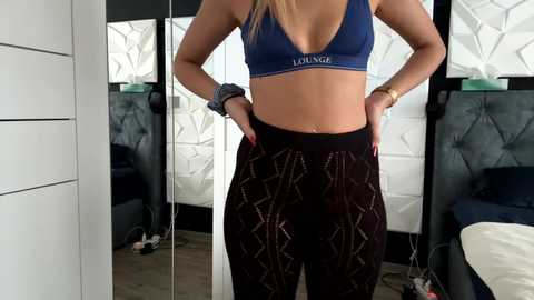 Media: Video of a fit, blonde woman in a blue sports bra and high-waisted, patterned leggings, standing with hands on hips in a modern bedroom with white, geometric-patterned wall art.