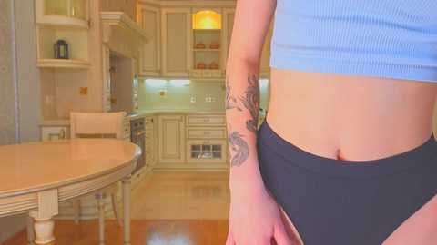 Media: Video of a woman's midriff in black panties, showing a floral tattoo on her left arm, standing in a beige kitchen with wooden floors, white cabinets, and a round table.