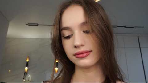 Media: Video of a young Caucasian woman with straight brown hair and fair skin, wearing a light-colored top, standing indoors with modern, minimalist decor in the background.