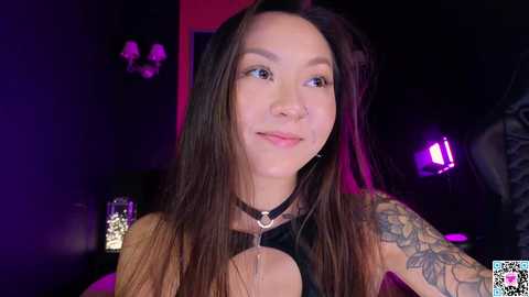 Media: Video of a young Asian woman with long black hair and a floral tattoo on her left arm, wearing a black choker, smiling in a dimly lit room with purple and pink lighting.