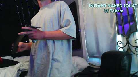 Media: A video shows a man in a loose, light blue t-shirt, holding a tablet, in a dimly lit room with a bed, a window with sheer curtains, and a black and purple light. Text reads, \"INSTANT NAKED SOUL 35 TUBES.\