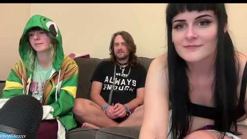 Media: Video of a young white woman with black hair, wearing a black bra, sitting on a couch next to two men in hoodies.