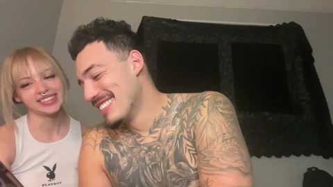 Media: Video of a smiling, light-skinned man with a tattooed chest and a blonde woman with bangs, both wearing casual attire, in a dimly lit room.