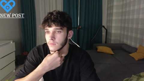 Media: Video of a young man with curly hair, wearing headphones, sitting on a grey couch, in a dimly lit room with a white dresser and teal curtains.