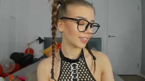 Media: Video of a young woman with fair skin, brown hair in two braids, wearing black glasses and a black fishnet top. Background includes a white wall, desk, and a lamp.