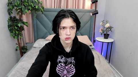 Media: Video of a pale-skinned, androgynous person with shoulder-length black hair, wearing a black hoodie with a white skull design, sitting on a bed with pink curtains and a green headboard. The room features a small table with a vase of flowers and a tall potted plant.