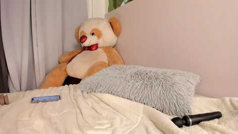 Media: Video of a teddy bear with red nose and collar, lying on a white bed with a light blue pillow and a vibrator beside it, in a room with light-colored walls and a window.