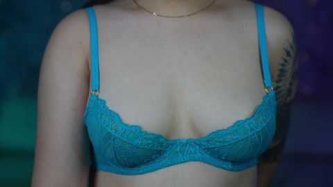 Media: Video of a woman's upper torso in a blue lace bra, showcasing fair skin and a tattoo on her left shoulder. She wears a thin gold necklace. Background is blurred, possibly green and purple hues.