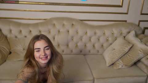 Media: Video of a young woman with long, wavy brown hair, wearing a beige sweater, smiling on a plush, cream-colored tufted sofa with gold accents and decorative pillows.