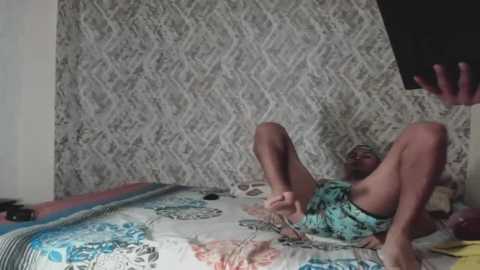 Media: Video of a light-skinned woman with short brown hair lying on a bed with patterned sheets, wearing a turquoise floral dress, in a dimly lit room with a white, patterned wallpaper.