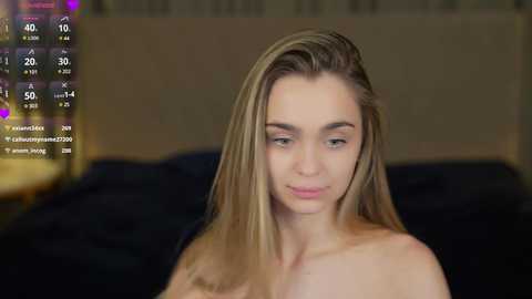 Media: Video of a topless, fair-skinned young woman with long blonde hair, looking slightly downcast, in a dimly lit bedroom with a dark bed and a beige headboard.