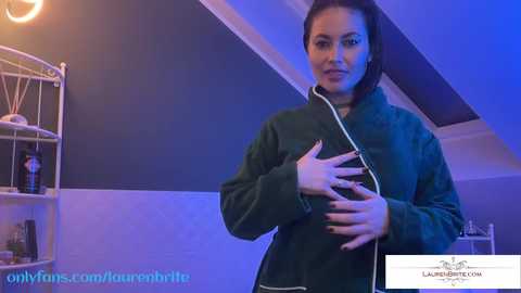 Media: Video of a dark-haired woman with a medium build, wearing a dark green jacket, standing indoors, hands on her chest, under blue lighting, OnlyFans watermark in the corner.