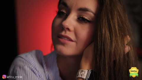 Media: Video of a young woman with fair skin, brown hair, and light makeup, wearing a light blue sweater, against a red background. She has a calm, contemplative expression.