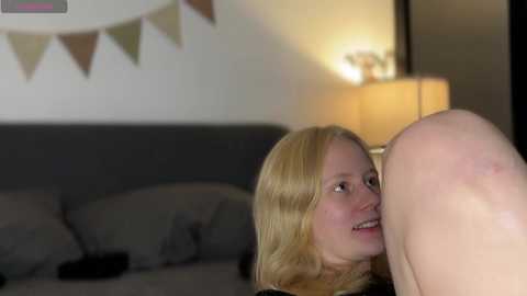 Media: Video of a blonde woman with fair skin and a light complexion, smiling, in a dimly lit bedroom with a grey bed and a string of bunting.