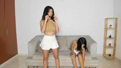 Media: Video of two young women with medium skin tones, one standing, the other kneeling, in a minimalist living room with light beige couch and wooden shelves.