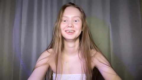 Media: Video of a young, fair-skinned girl with long, straight blonde hair smiling broadly. She wears a white towel and stands against a blurred, light-colored curtain background.