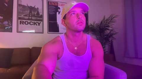 Media: Video of a shirtless, muscular man in a white tank top, wearing a yellow baseball cap, sitting on a couch under purple lighting. Behind him are framed movie posters and a potted plant.