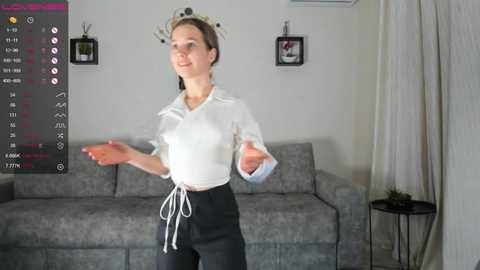 Media: Video of a woman in a white blouse, black pants, and a floral headband, dancing in a modern living room with a gray couch, framed artwork, and a black side table.