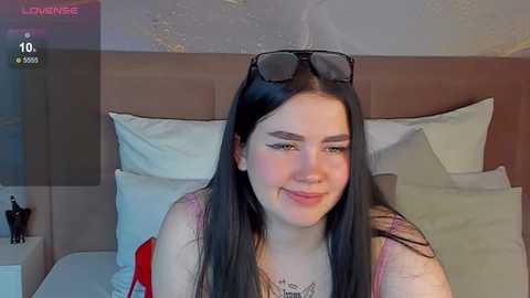 Media: Video of a young woman with long black hair, wearing black sunglasses, lying on a bed with white pillows and a brown headboard. She has fair skin, a slender build, and a slight smile.