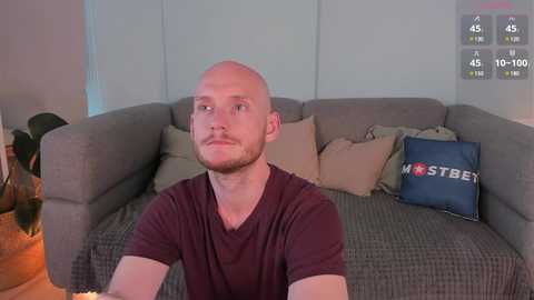 Media: Video of a bald, fair-skinned man with a beard, wearing a maroon T-shirt, sitting on a gray couch with beige cushions. The background shows a gray wall and a \"MOSTBET\" pillow.