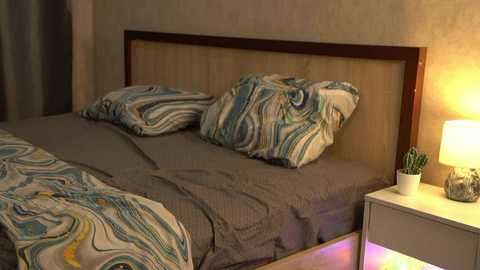 Media: Video of a modern bedroom with a bed featuring a wooden headboard, a gray comforter, and two pillows with a wavy, abstract pattern. The nightstand has a lamp, a small plant, and a vase with pencils.