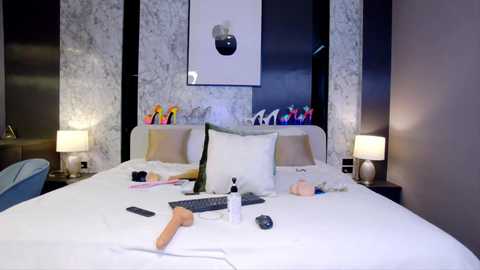 Media: Video of a luxurious, modern hotel room with a white bed, white pillows, black-and-white marble wallpaper, two bedside lamps, colorful pens, and a sex toy on the bed.