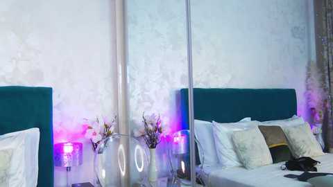 Media: Video of a modern, minimalist bedroom with two teal upholstered beds, white linens, and a silver vase with flowers on a mirrored nightstand. Soft, ambient purple lighting creates a serene atmosphere.