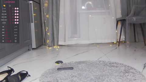 Media: A video of a modern living room with light grey walls, a white tiled floor, and a plush grey rug. A smartphone lies on the rug, and a pair of black shoes is nearby. The room is softly lit by fairy lights, with a glass door in the background.