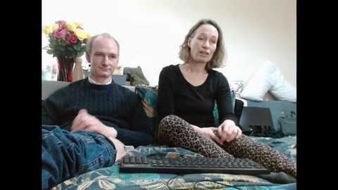 Media: Video of a bald, middle-aged man and a blonde woman in leopard-print leggings sitting on a patterned couch in a cluttered living room.