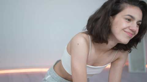 Media: Video of a young Caucasian woman with shoulder-length dark hair, wearing a white spaghetti-strap tank top and gray sweatpants, smiling softly. Background is a light gray wall and wooden floor.