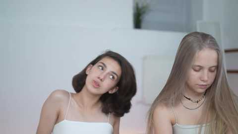 Media: Video of two young women, one with shoulder-length dark hair and the other with long blonde hair, wearing tank tops, looking thoughtful in a minimalist, light-colored room.