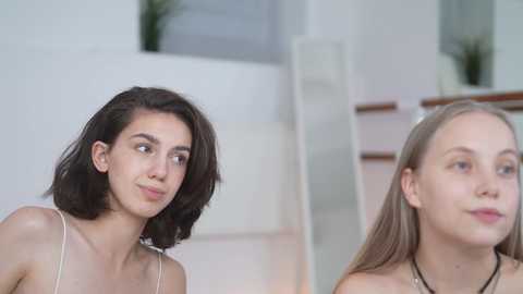 Media: Video of two young women with fair skin, shoulder-length hair, and slender physiques. One with dark hair wears a white tank top, the other with light brown hair wears a black necklace. They stand in a modern, minimalist room with white walls and a wooden staircase.