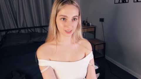 Media: Video of a young, fair-skinned woman with long blonde hair, wearing an off-the-shoulder white top, standing in a dimly lit bedroom with grey curtains, black bed, wooden nightstand, and a lamp.