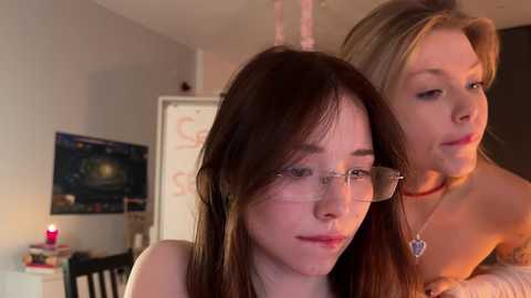 Media: Video of two young women with fair skin and long brown hair; one with glasses, wearing a black top, the other in a pink top. They stand in a cozy, dimly lit room with a TV, a chalkboard, and a wooden chair.