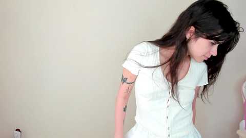 Media: A video of a young woman with long dark hair, wearing a white button-up shirt, leaning against a plain beige wall. Her left arm has a tattoo, and her expression is contemplative.