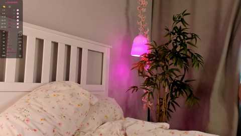 Media: Video of a cozy bedroom with a white wooden bed, floral-patterned bedding, a tall potted plant, and a pink lampshade casting a soft glow.