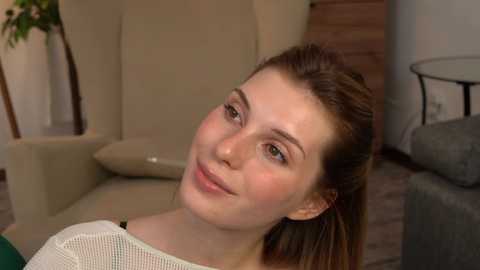 Media: A video of a fair-skinned woman with light brown hair in a ponytail, smiling, wearing a white mesh top, sitting in a beige chair in a modern living room with wooden floors and a gray sofa.