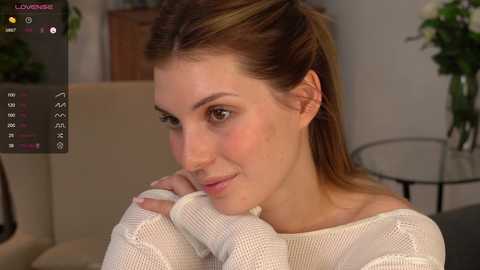 Media: Video of a fair-skinned woman with straight, shoulder-length brown hair, wearing a white knitted sweater, leaning forward with her chin resting on her hands, smiling softly. Background includes a beige sofa and green plants.