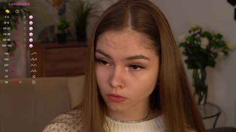 Media: Video of a young woman with light skin and straight, medium-brown hair, wearing a white sweater, with a pensive expression, sitting in a cozy, dimly lit room with a potted plant and a wooden dresser in the background.