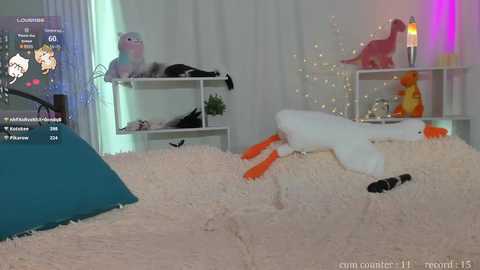 Media: A video of a plush duck and dog on a white shag rug, with a teal pillow in the background. The room features white shelves and a pink dinosaur toy, with fairy lights and a window.