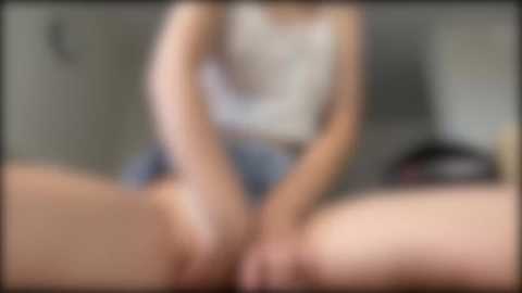 Media: Blurry video of a woman in a tank top and shorts, legs spread, sitting on a bed, with a blurred background.