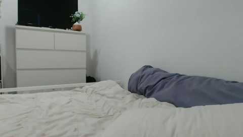 Media: Video of a minimalist bedroom with a white dresser, a flat-screen TV, and a neatly made bed with a light blue duvet. The room has a clean, modern aesthetic with soft lighting.