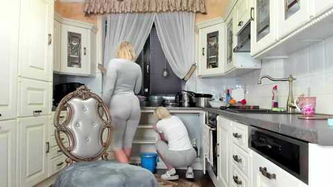 Media: Video of a modern, white kitchen with a woman in grey workout attire and a blonde in a white shirt scrubbing the floor.