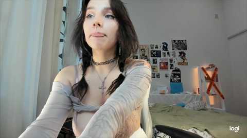 Media: A young woman with long dark hair, wearing a choker, braids, and a sheer top, stands in a cluttered bedroom adorned with posters and a bed with white sheets.