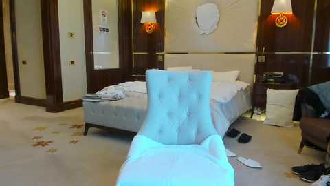 Media: Video of a luxurious, modern hotel room with a tufted, light blue chaise lounge, beige carpet, a king-sized bed, and a round wall clock.